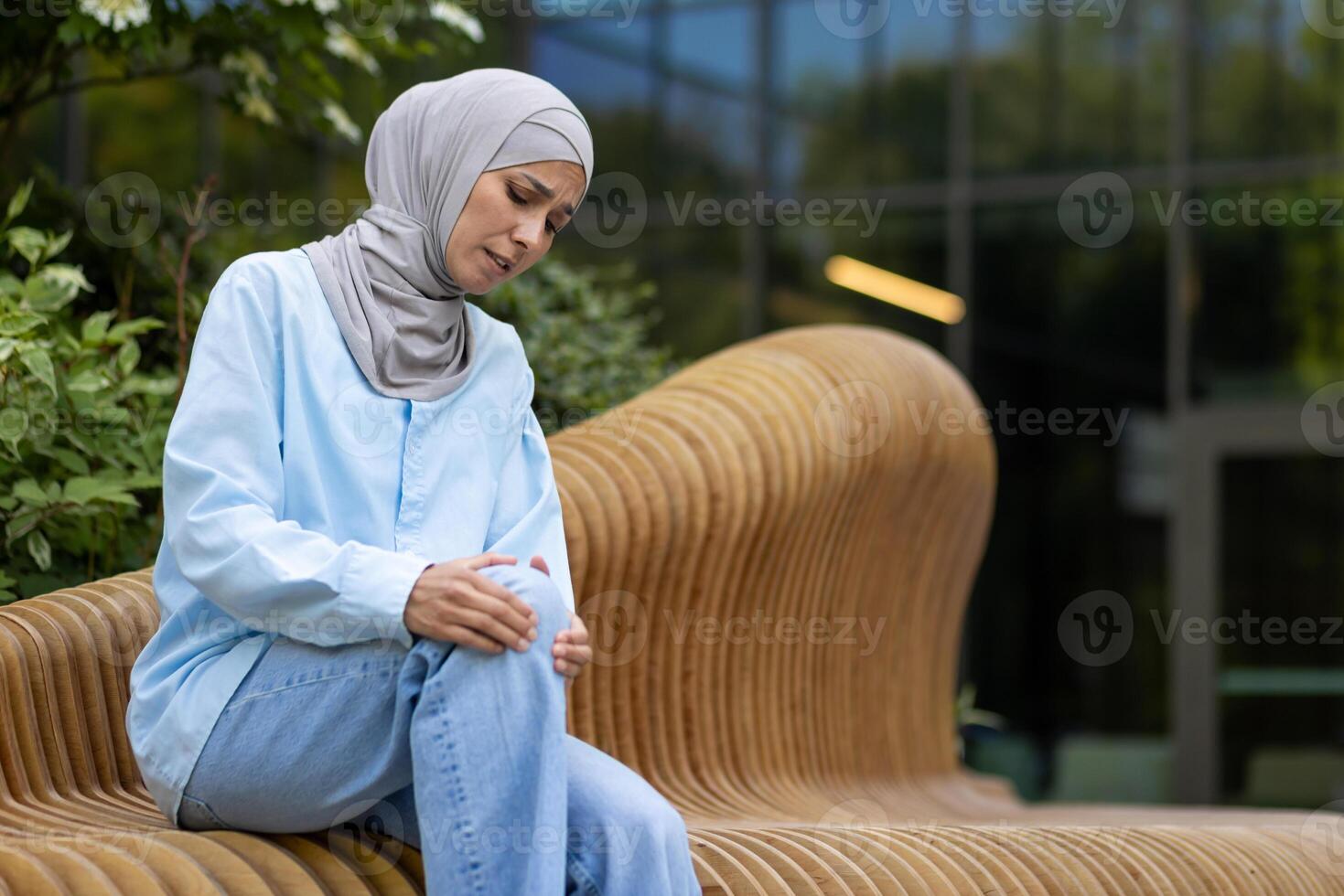 Muslim weiblich Sitzung draußen und erziehen Bein gebogen im Knie mit Leiden Ausdruck auf Hintergrund von modern Gebäude. krank Frau gegenüber Trauma Konsequenzen und Reiben verletzt Platz mit beide Hände. foto