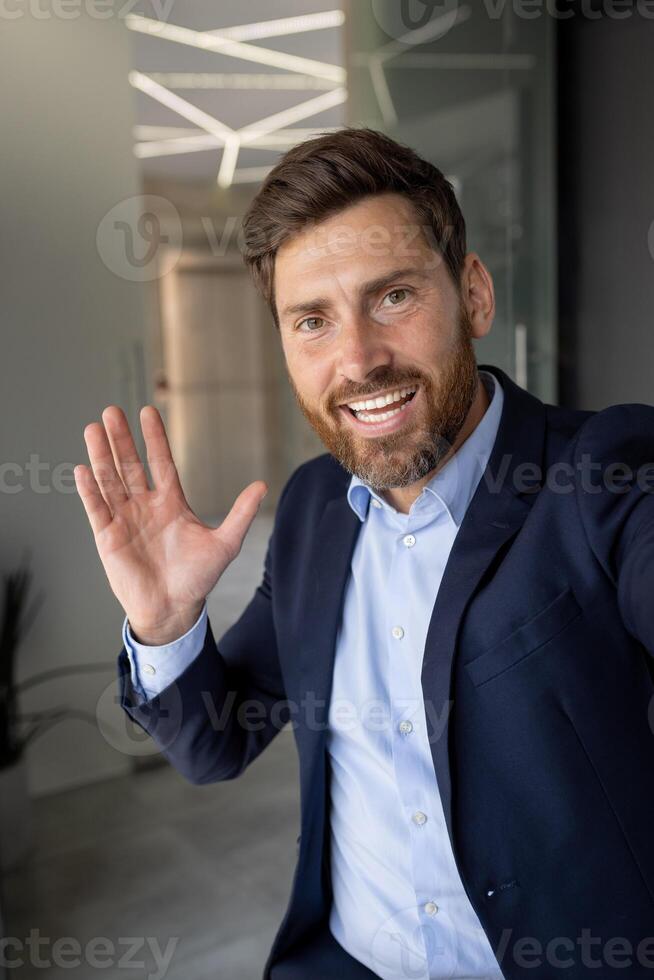 Porträt von ein jung lächelnd Geschäftsmann, Makler, Verkäufer Stehen im das Büro reden auf das Kamera Telefon, lächelnd und gestikulieren mit seine Hände. Nahansicht Vertikale Foto. foto