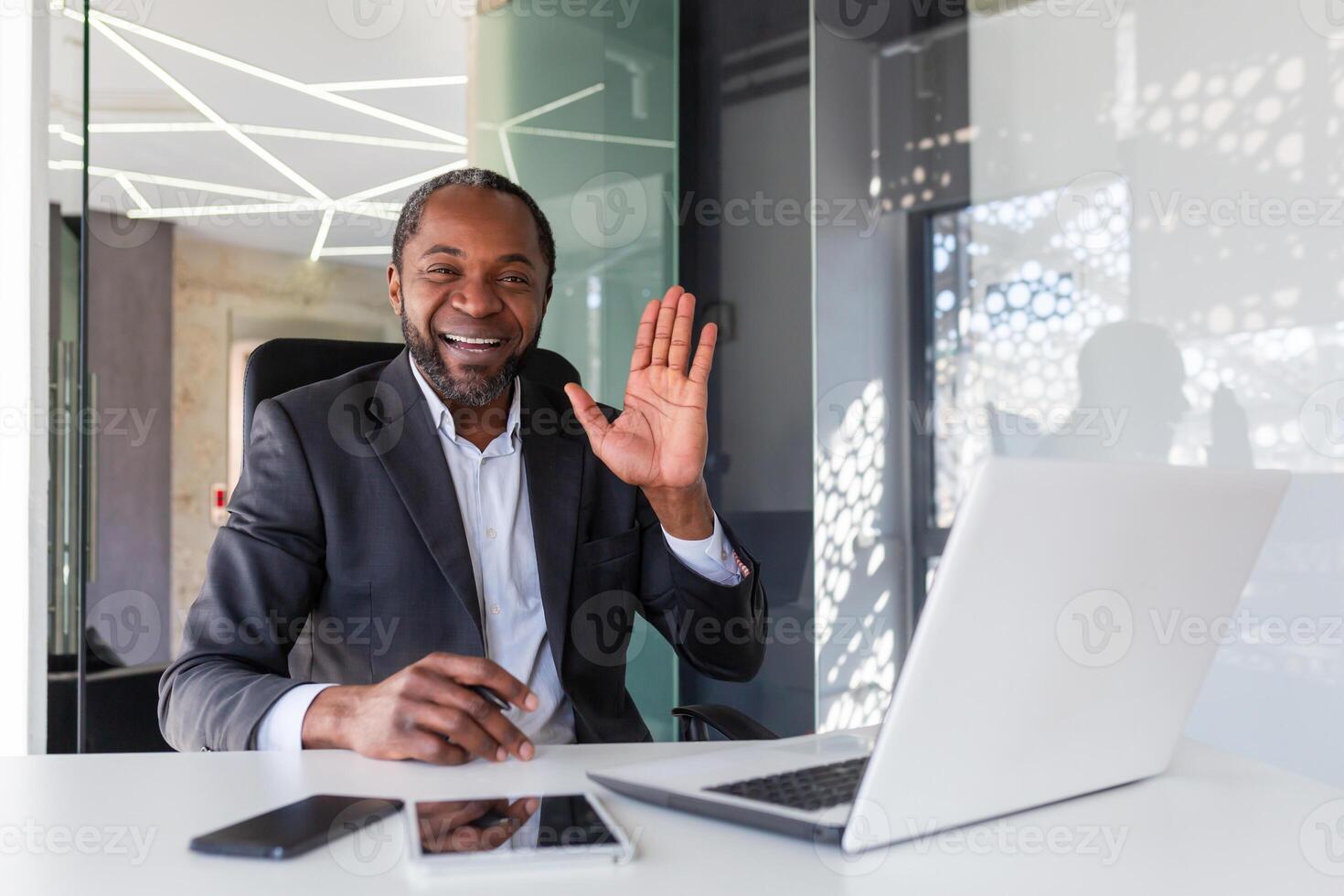 Porträt von erfolgreich reifen afrikanisch amerikanisch Chef, Mann lächelnd und suchen beim Kamera Sitzung beim Schreibtisch Innerhalb Büro, Geschäftsmann winken Hand Geste von Gruß und Freundschaft. foto