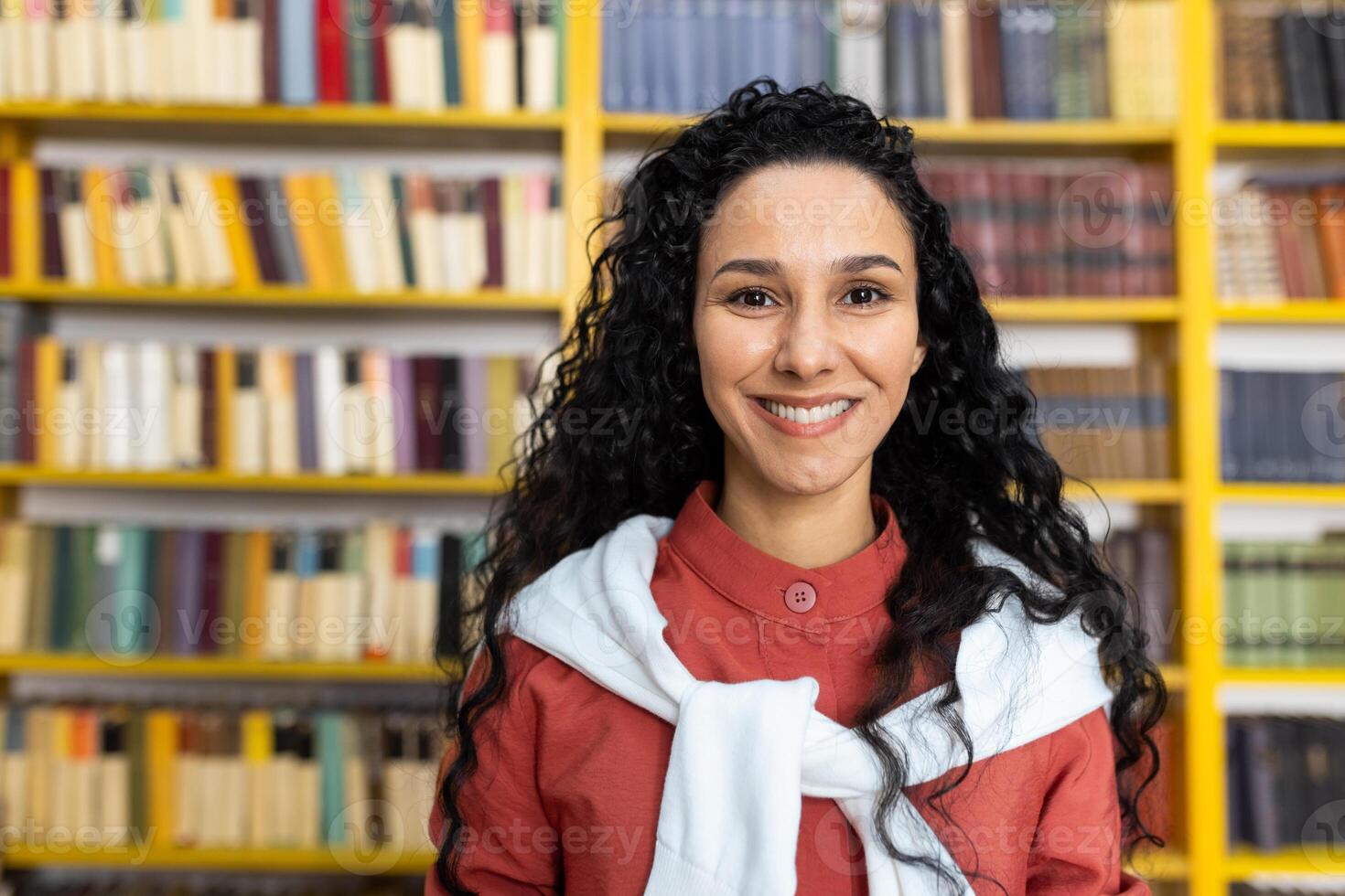 ein jung spanisch weiblich Schüler steht im Vorderseite von ein bunt Bücherregal im ein Bibliothek, symbolisieren Ausbildung, Wissen, und Diversität. foto