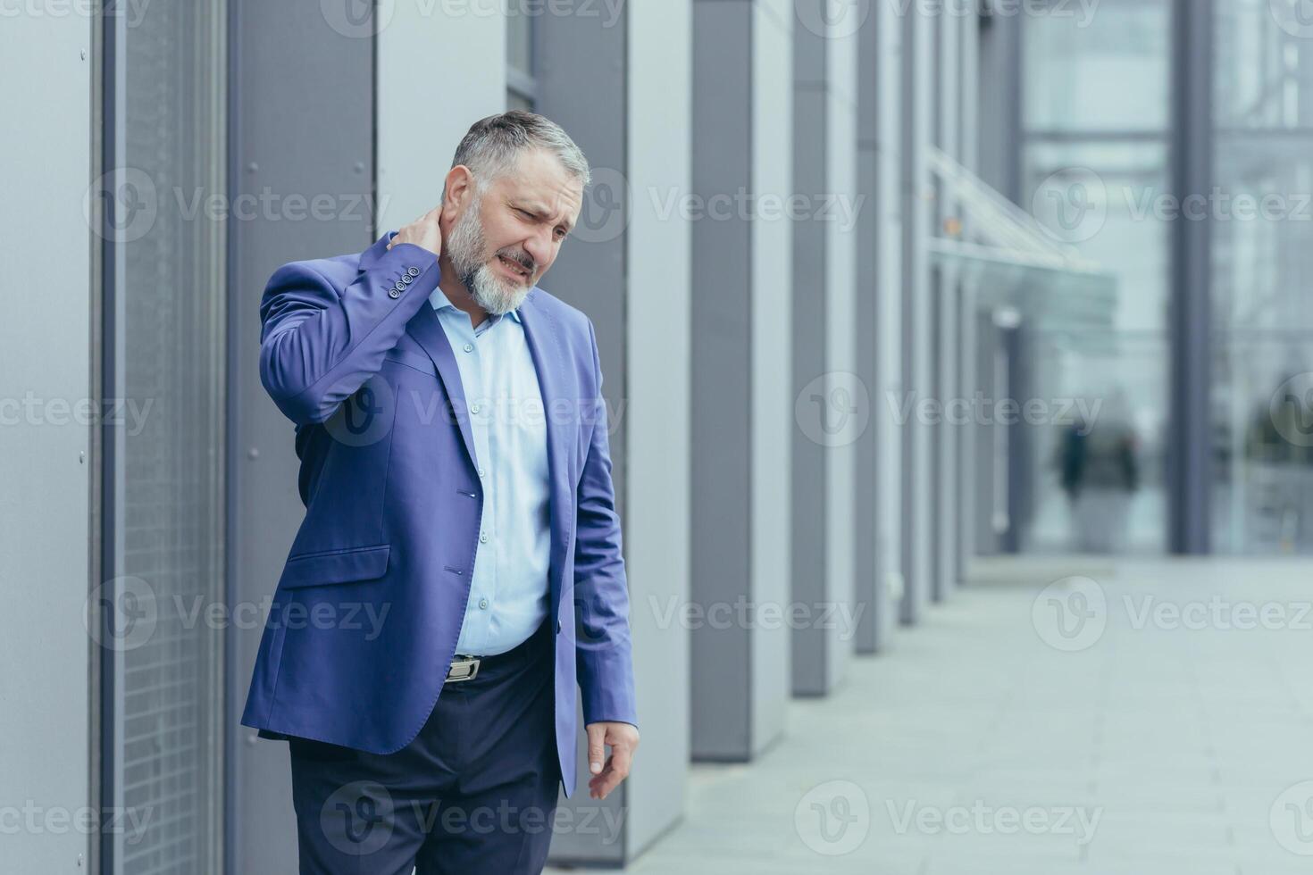 ein Senior Mann, ein Banker, ein Büro Arbeiter im Anzug, ging draußen das Büro zum ein Mittagessen brechen, seine Hals tut weh, er Massagen seine Hals mit einer Hand, er fühlt sich Schmerz. foto