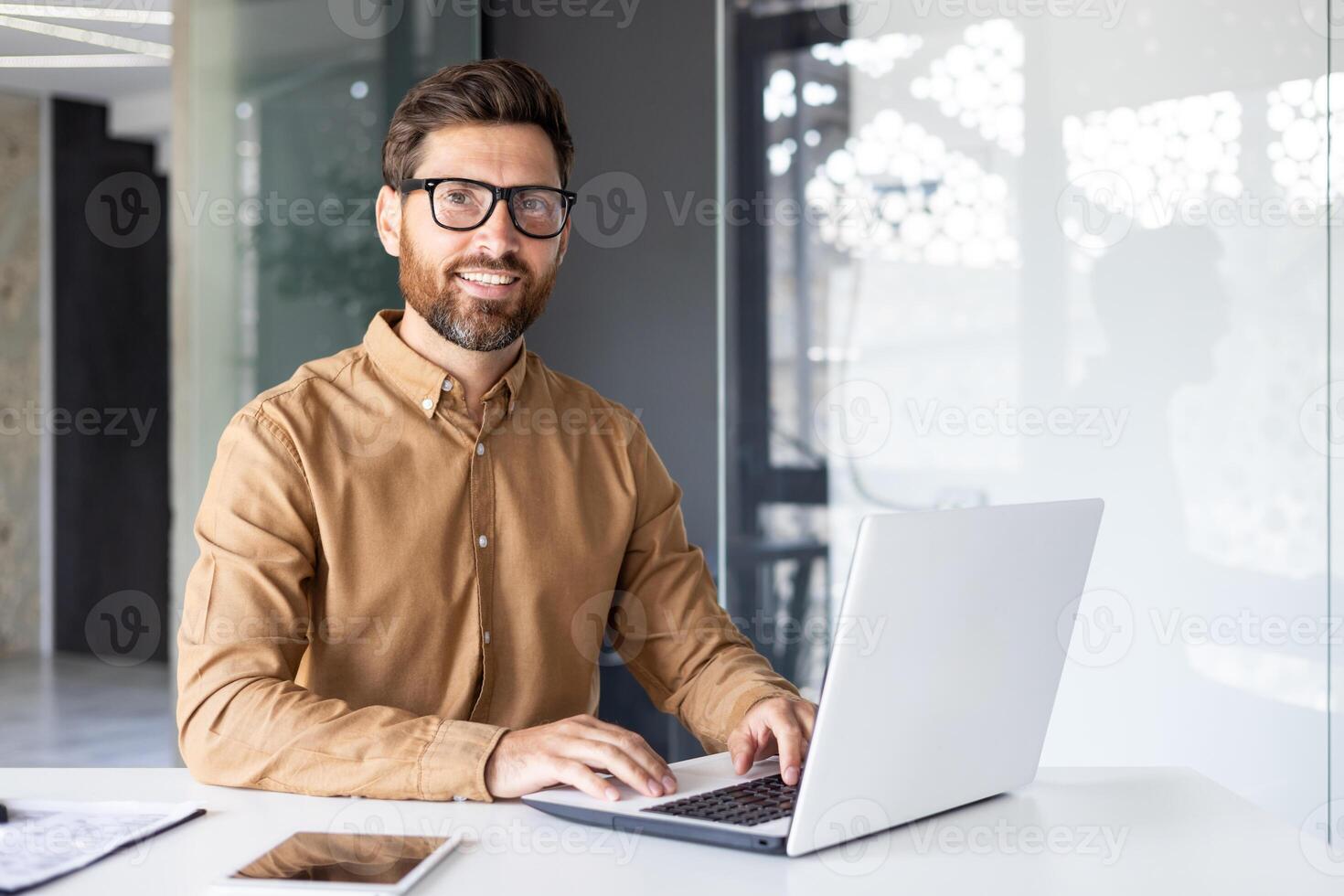 Porträt von erfolgreich Mann beim Arbeitsplatz Innerhalb Büro, erfahren lächelnd Geschäftsmann im Hemd lächelnd und suchen beim Kamera, Programmierer Entwicklung Neu Software mit Laptop. foto