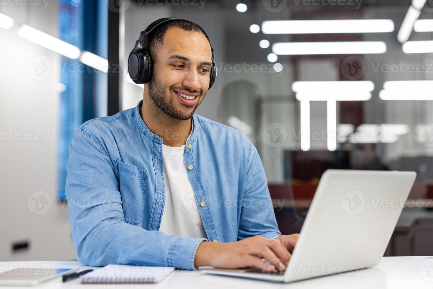 lächelnd indisch Mann im ein Zuhause Büro Einstellung, Arbeiten auf ein Laptop während tragen Kopfhörer im ein modern Leben Zimmer. foto