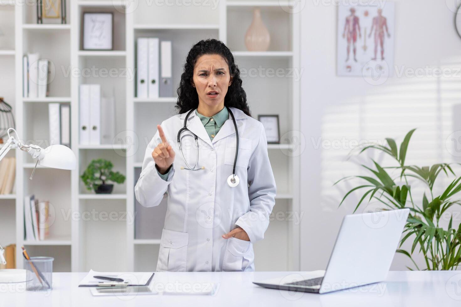 Latein amerikanisch jung weiblich Arzt Stehen im Klinik Büro in der Nähe von Arbeiten Schreibtisch, suchen Ernsthaft beim Kamera und zeigen Finger Verbot Geste. foto