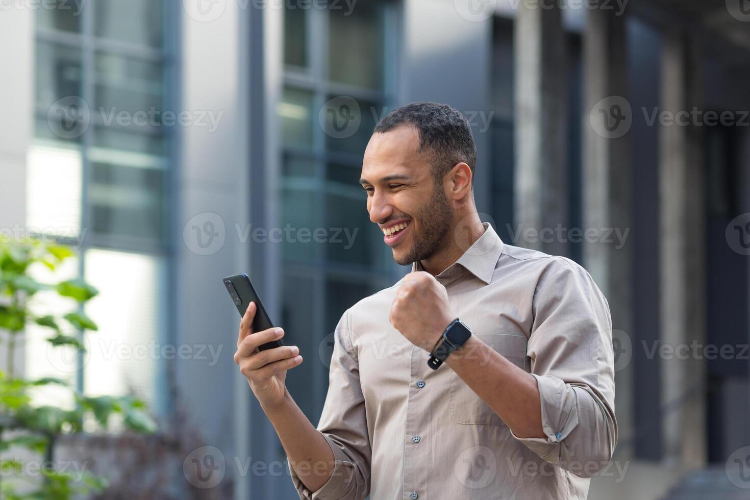 afrikanisch amerikanisch Geschäftsmann draußen Büro Gebäude mit Telefon, lächelnd und glücklich halten Hand oben Triumph Geste, jung Unternehmer feiern Sieg lesen gut Investition Nachrichten online foto