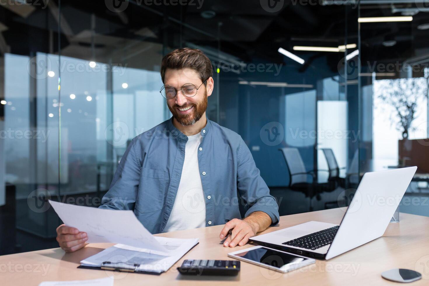 erfolgreich Financier Investor funktioniert mit Unterlagen und Konten Innerhalb Büro, Geschäftsmann mit Bart lächelnd zufrieden mit Arbeit Ergebnisse und Erfolge, Mann Innerhalb Büro mit Laptop Arbeiten. foto