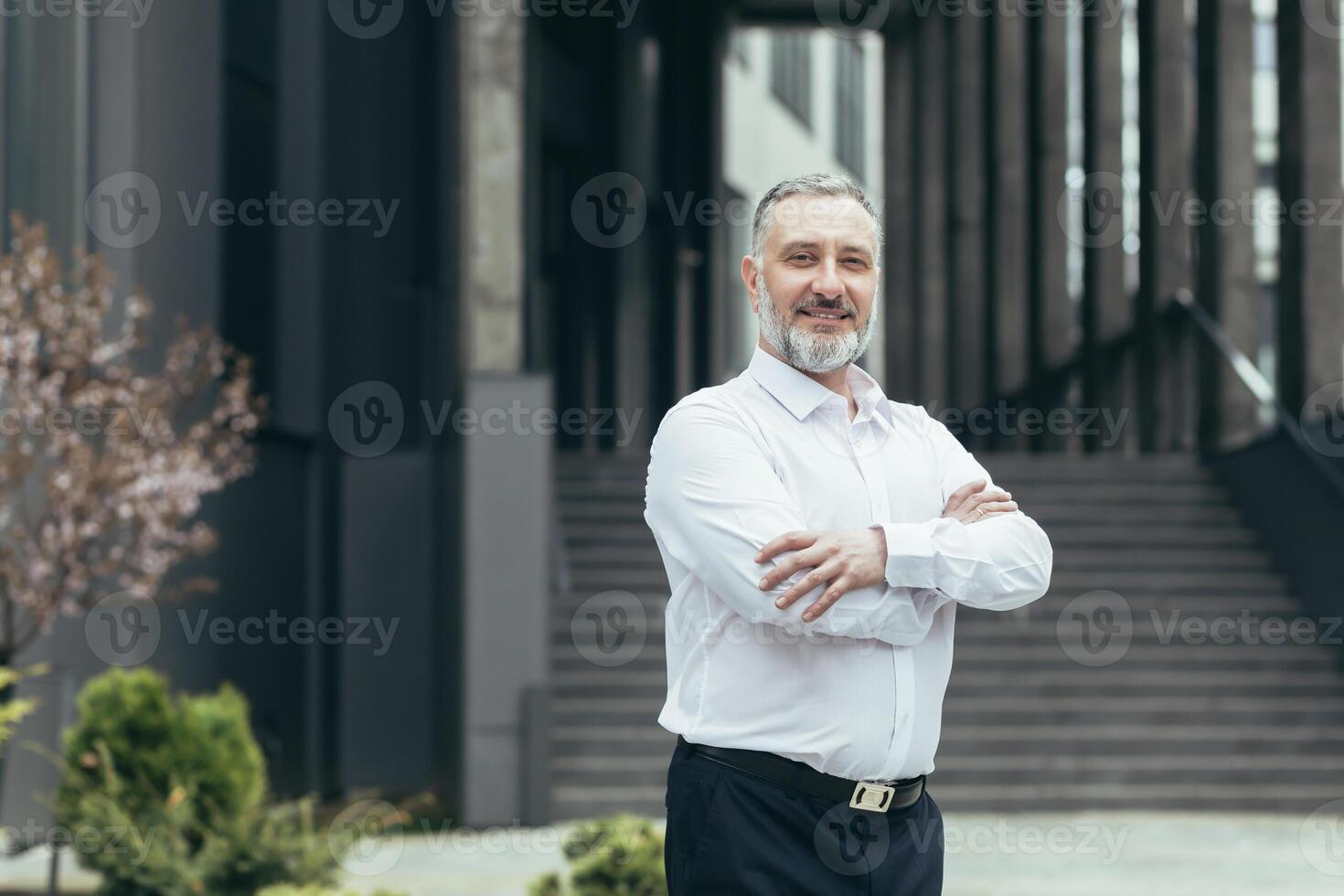 Porträt von ein Senior Mann, ein Lehrer, ein Dozent Stehen im ein Weiß Hemd gegen das Hintergrund von ein modern Gebäude von ein lehrreich Institution. lächelnd, aussehen beim das Kamera foto