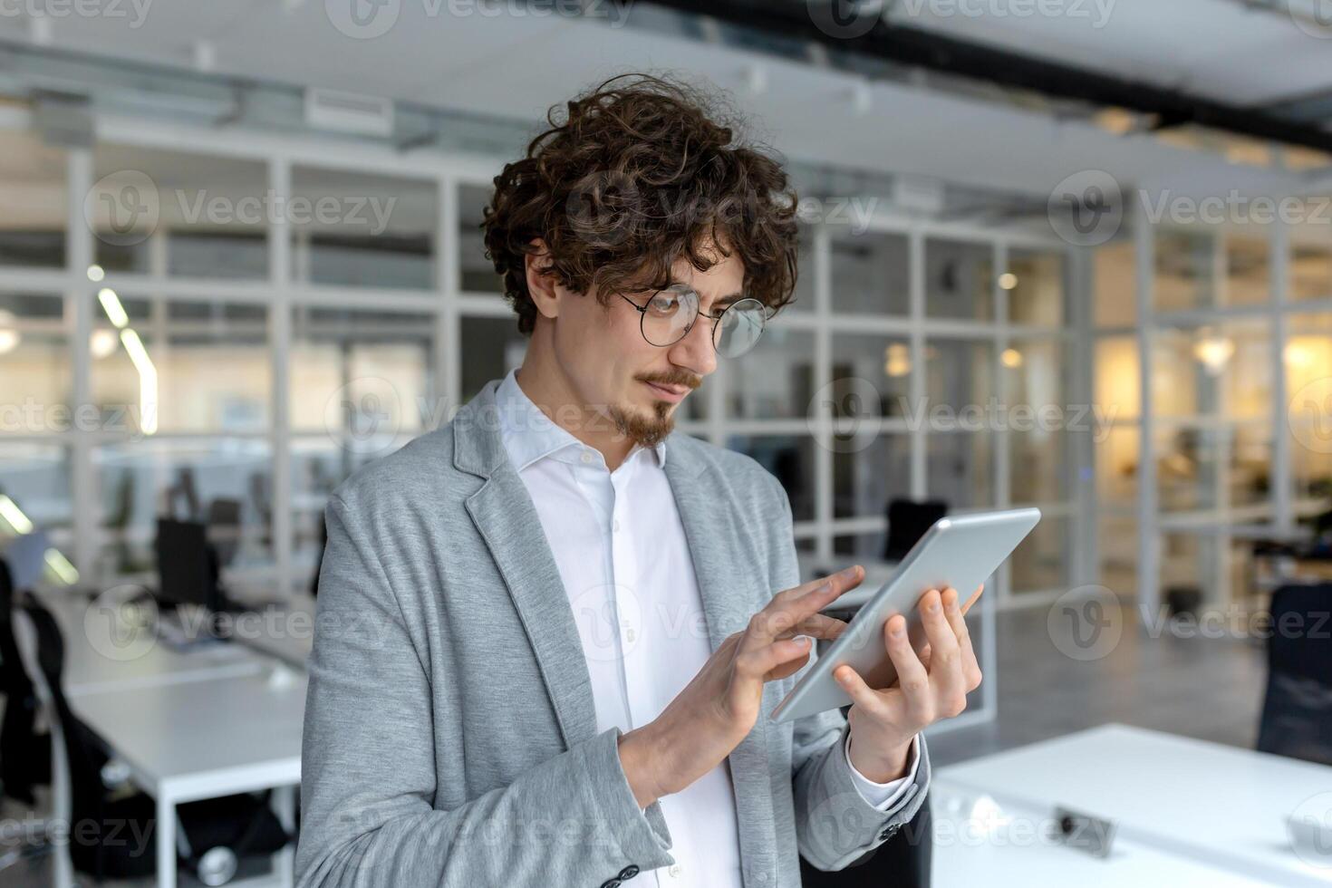 lockiges Haar Geschäftsmann im ein passen Jacke vertieft im seine Tablette Arbeit beim ein hell korporativ Büro. foto