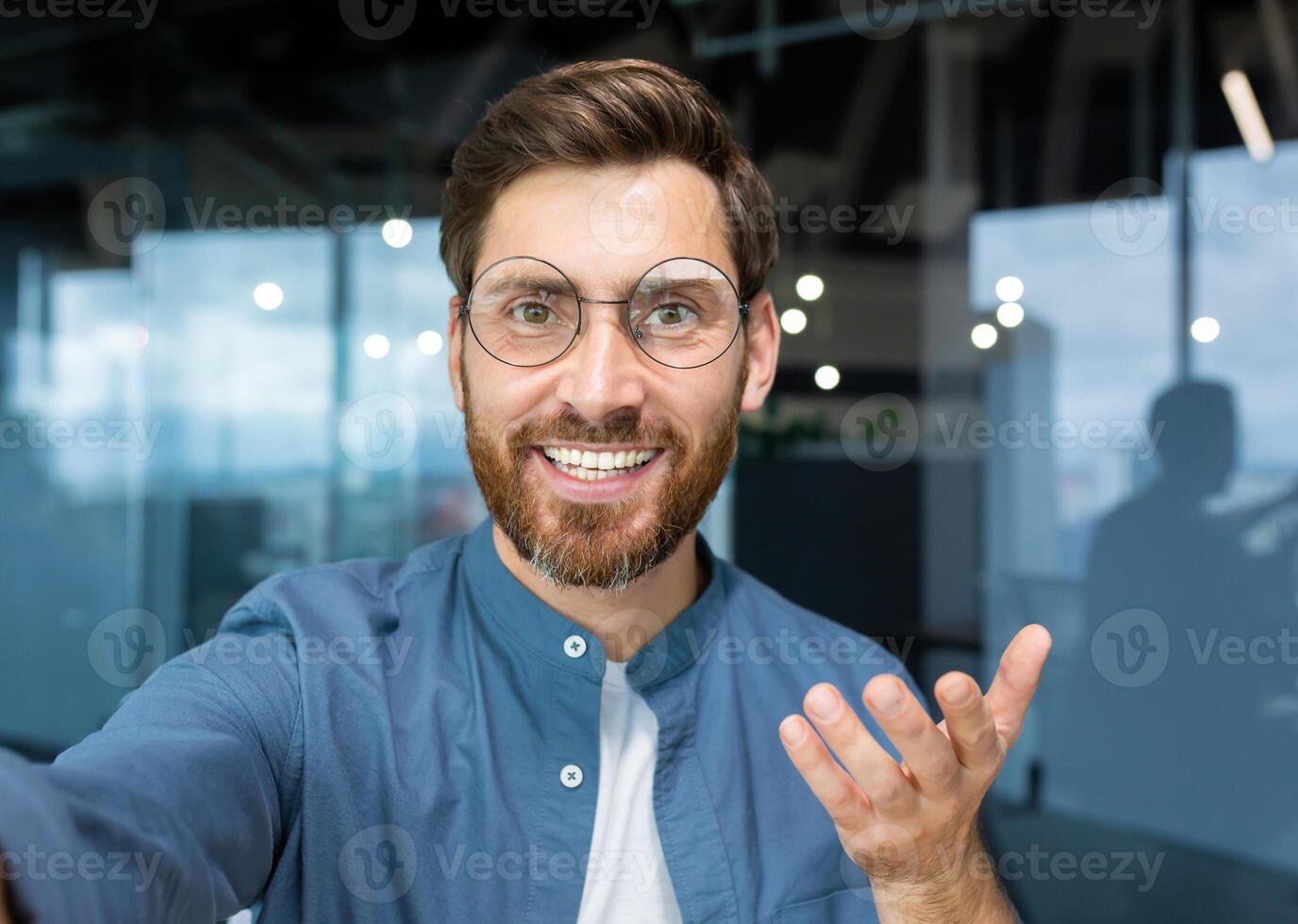 Porträt von erfolgreich Geschäftsmann im beiläufig Hemd und Brille Mann suchen beim Smartphone Kamera und lächelnd, Kamera Aussicht Anruf reden mit freunde Innerhalb Büro entfernt. foto