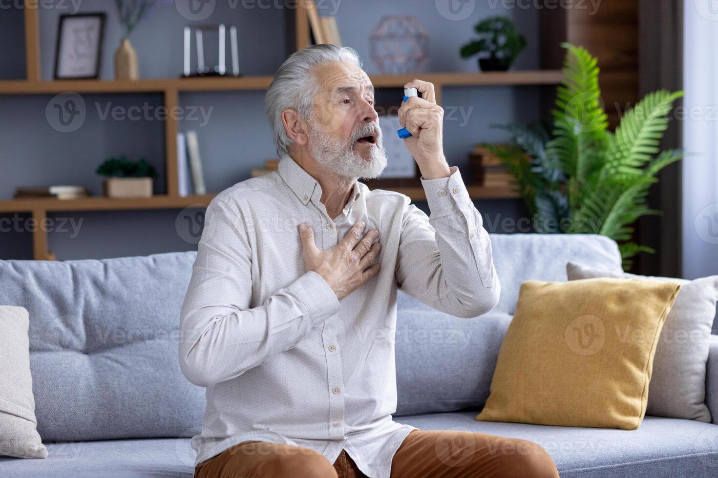 Alten kaukasisch Mann erleben ein Asthma Attacke, mit ein Blau Inhalator während Sitzung auf ein Couch im ein modern Leben Zimmer. foto