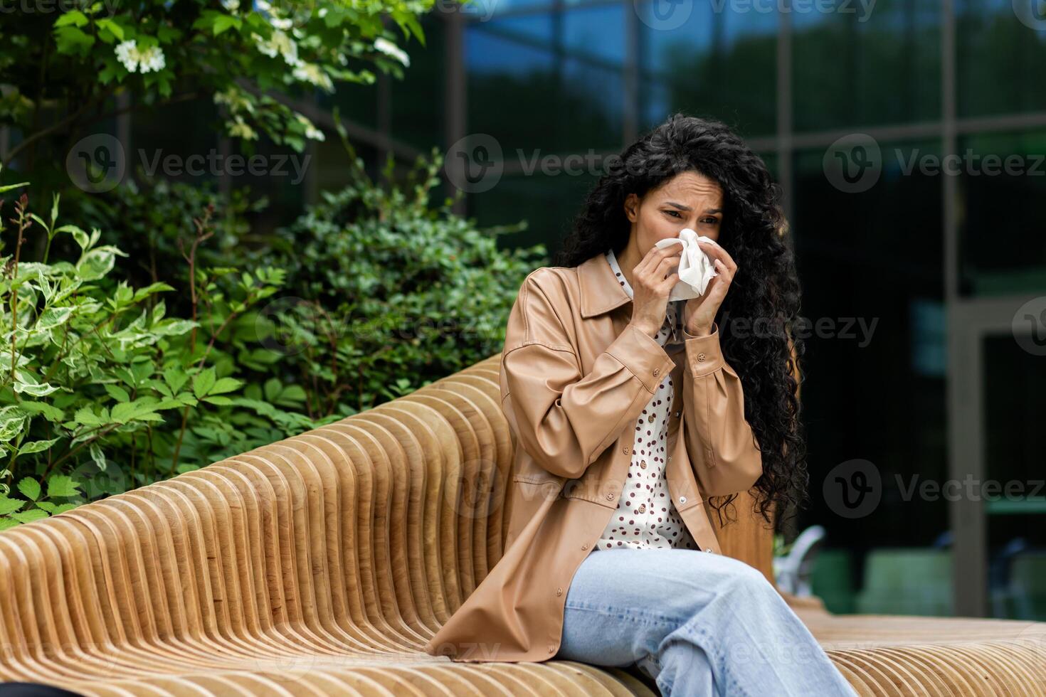 ein Frau sitzt auf ein draussen Bank, weht ihr Nase mit ein Gewebe, möglicherweise Leiden von kalt oder Allergien, im ein natürlich, städtisch Einstellung. foto