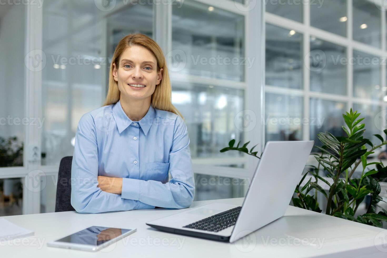 zuversichtlich weiblich Fachmann lächelnd beim Arbeitsplatz mit Laptop, Tablette, und Innen- Pflanze im ein zeitgenössisch Büro Einstellung. foto