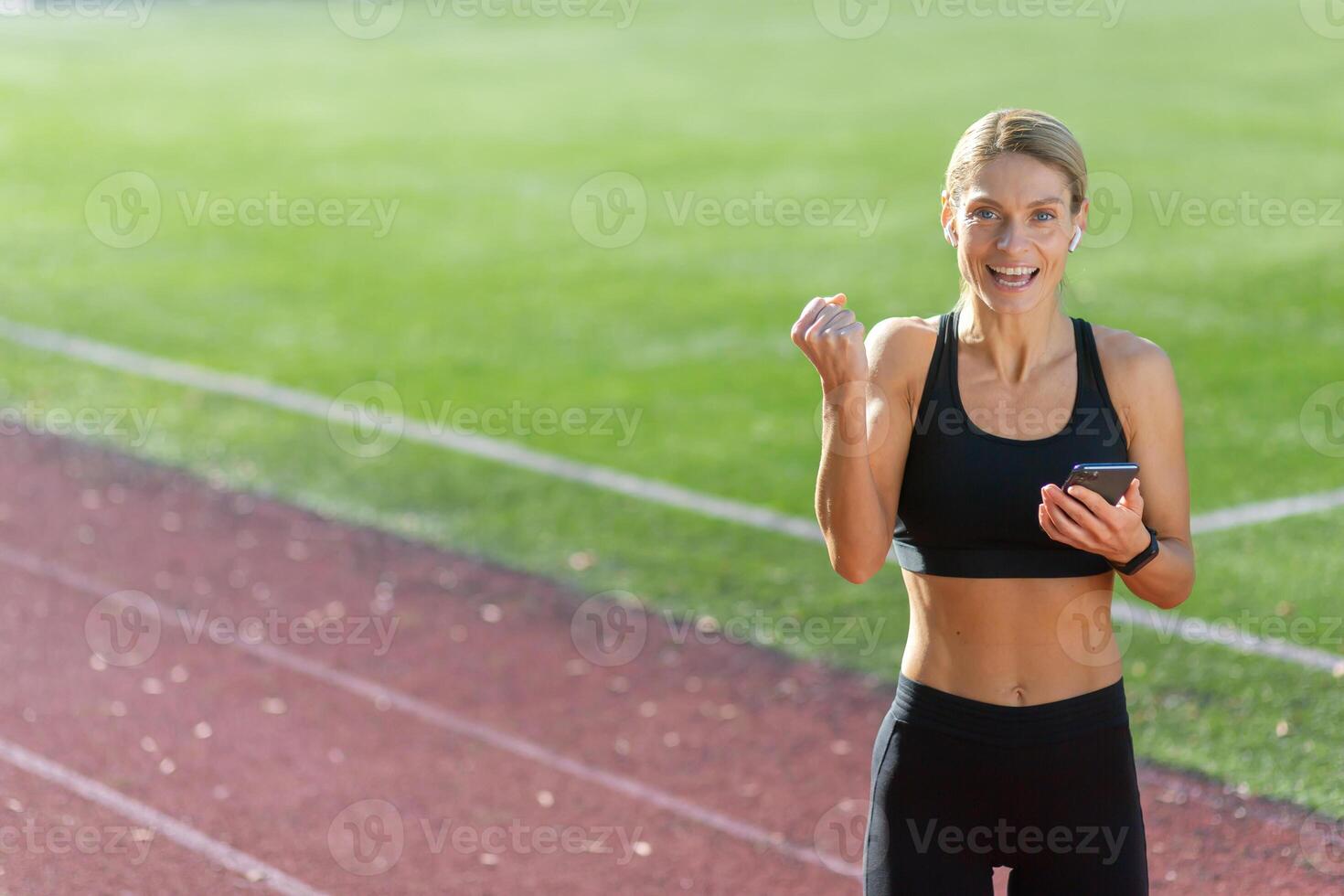 lächelnd sportlich Frau ruhen nach ein laufen, halten ihr Telefon und Auswahl Musik- beim das Stadion Spur auf ein sonnig Tag. foto