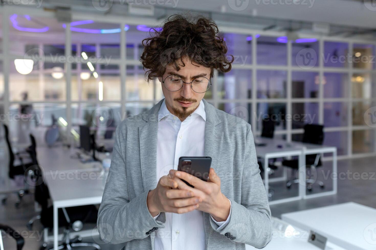 ernst männlich Unternehmer mit lockig Haar absorbiert im lesen ein Text Botschaft auf seine Telefon beim ein korporativ Büro. foto