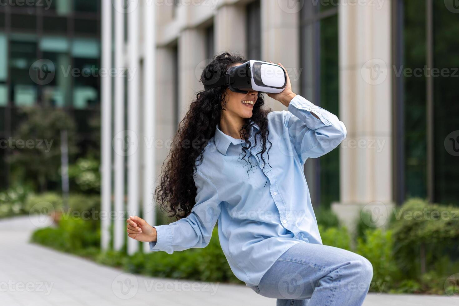 jung schön Mädchen im Hemd und Jeans Tanzen draußen Büro Gebäude tragen virtuell Wirklichkeit Gläser, spanisch Frau glücklich haben Spaß und entspannend mit lockig Haar. foto