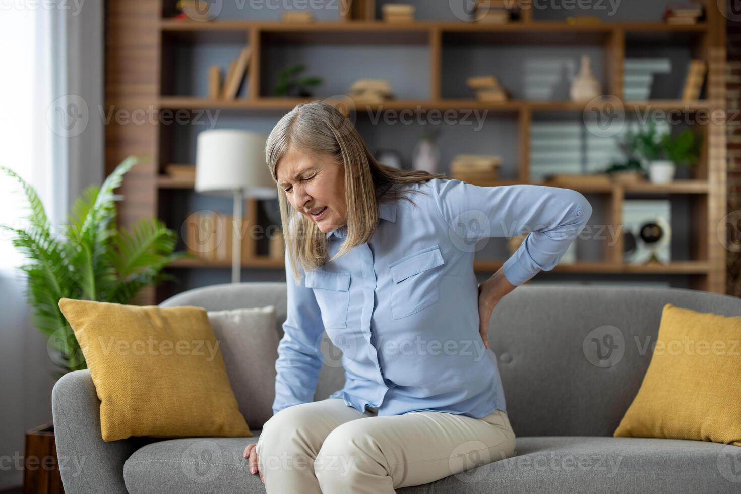 reifen Dame Gefühl Wirbelsäule Muskeln Schmerzen Attacke und halten zurück während Schielen Augen auf Sofa mit Kissen. krank Frau haben Problem mit Stehen oben und erleben Niere schmerzen im Leben Zimmer. foto