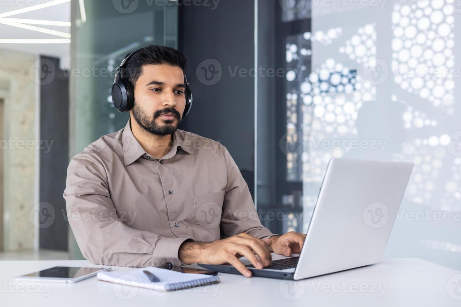 konzentriert Latino männlich im formal Hemd tragen modern Headset und mit tragbar Computer während Arbeiten durch Schreibtisch. zeitgenössisch Programmierer bekommen bereit zum Treffen mit Fachmann Garnitur. foto