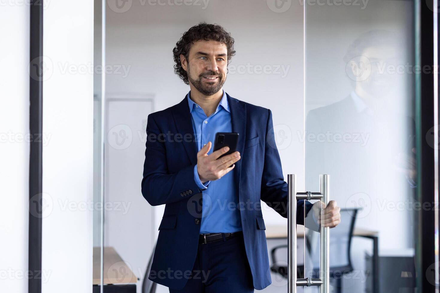 erfolgreich froh Geschäftsmann öffnet Glas Büro Tür, Mann mit Telefon im Hände lächelnd, zufrieden mit Leistung Ergebnisse, Boss im Geschäft Anzug. foto