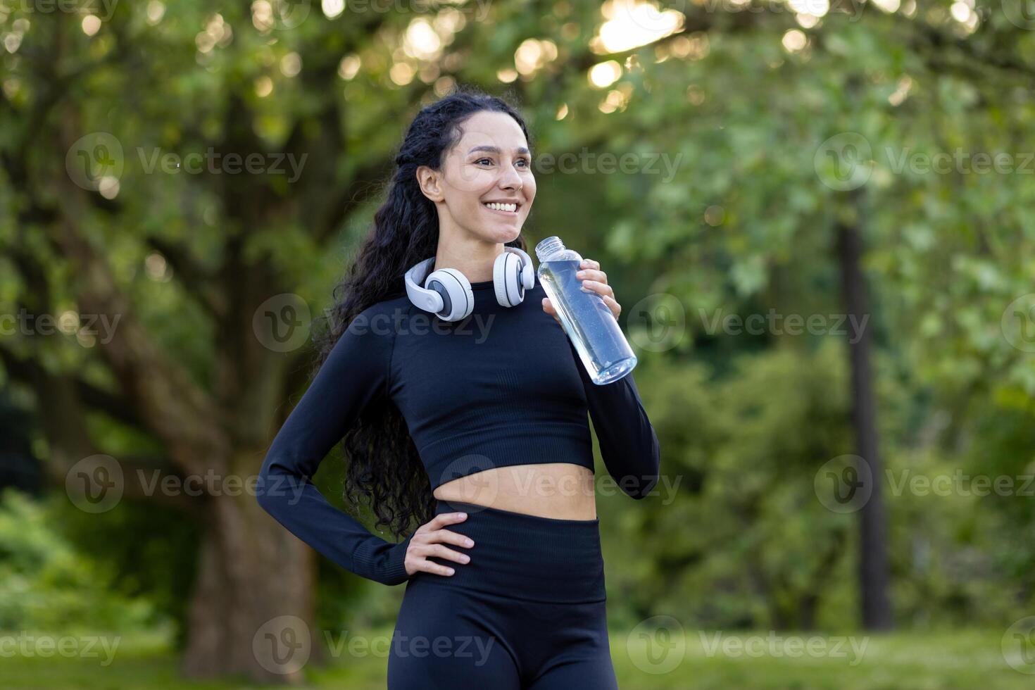 lächelnd aktiv Frau im trainieren Ausrüstung nehmen ein brechen mit ein Wasser Flasche im ein Grün Park Einstellung, Kopfhörer um ihr Nacken. foto