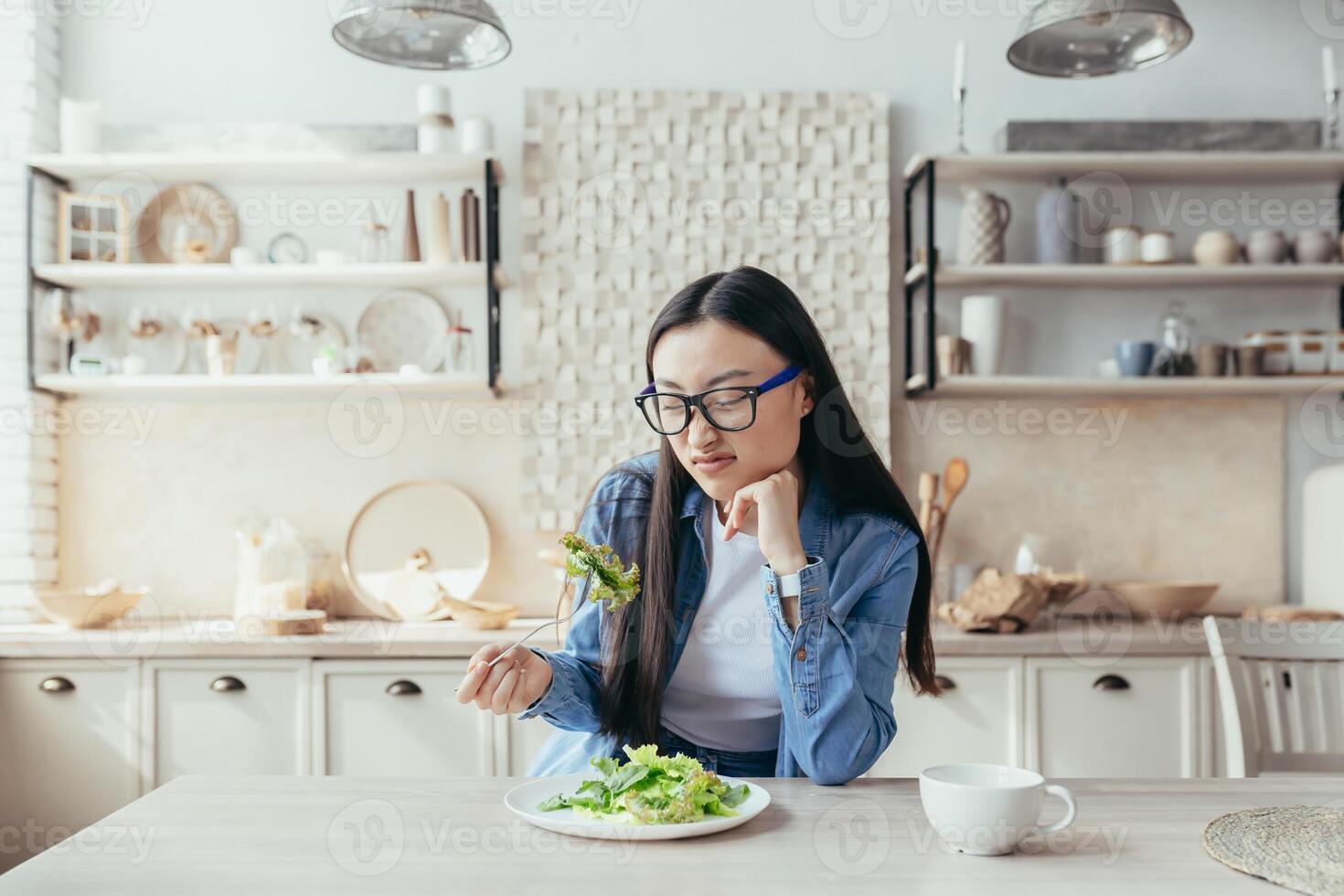 jung asiatisch Frau unzufrieden mit Diät im Küche, Frau Essen gesund Essen, versuchen zu Essen Salat und gesund Essen, Teenager beim heim. foto
