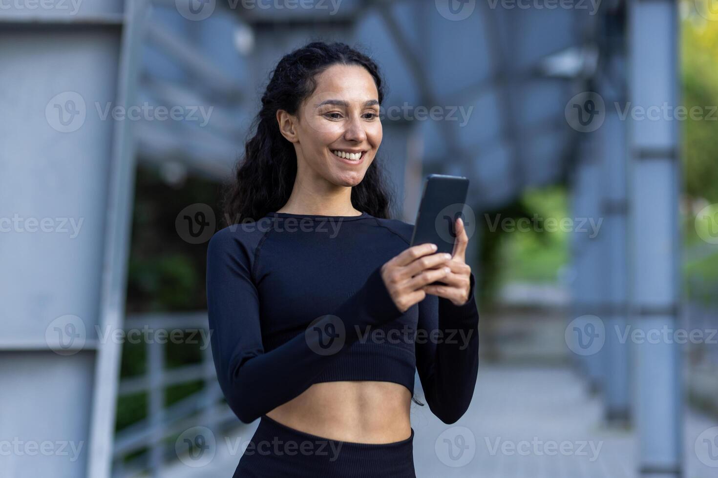 jung schön Frau mit Telefon, Sportlerin lächelnd und ruhen nach Laufen und tun aktiv physisch Übungen, spanisch Frau mit lockig Haar im Trainingsanzug. foto