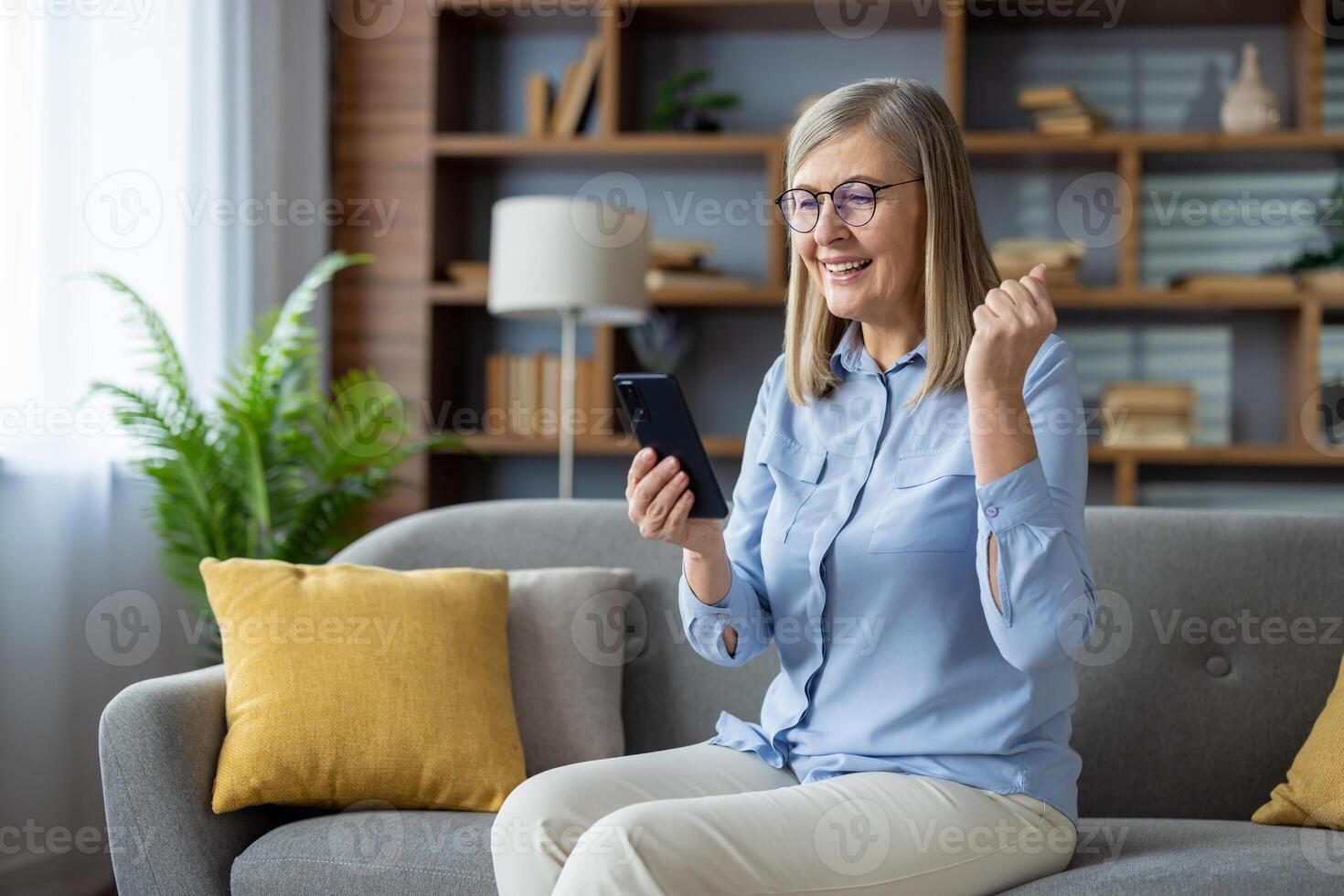 ein Senior Frau auf das Sofa Prost mit ein geballt Faust, suchen beim ihr Smartphone, verkörpern Freude und Erfolg. foto