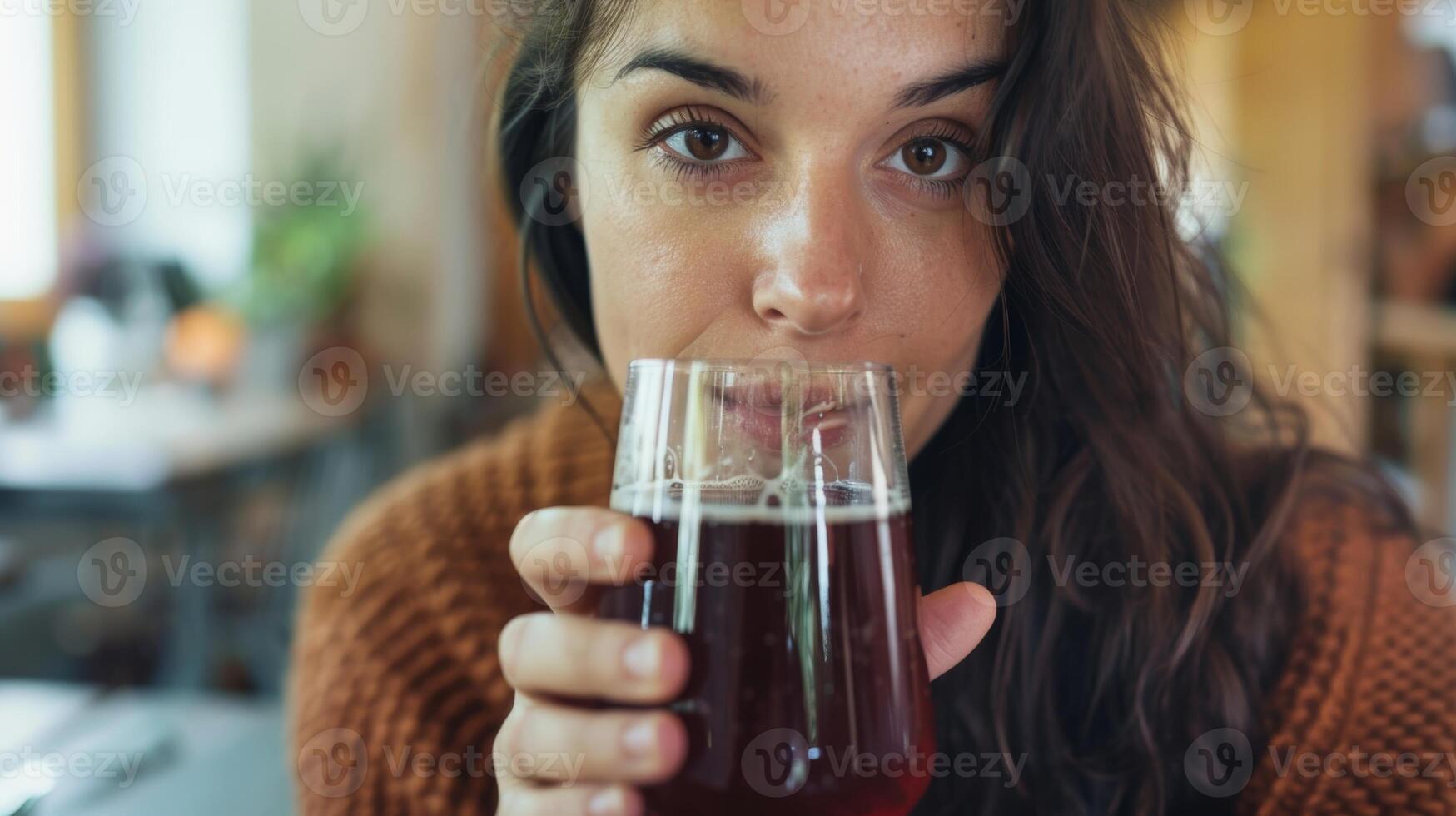 ein Frau hält oben ein Glas von dunkel rötlich-braun Kombucha ihr Gesicht zerknittert im Erwartung von das Geschmack foto