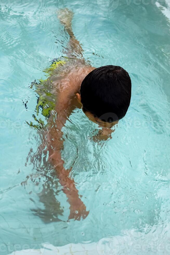 glücklich indisch Junge Schwimmen im ein Schwimmbad, Kind tragen Schwimmen Kostüm entlang mit Luft Tube während heiß Sommer- Urlaube, Kinder Junge im groß Schwimmen Schwimmbad. foto