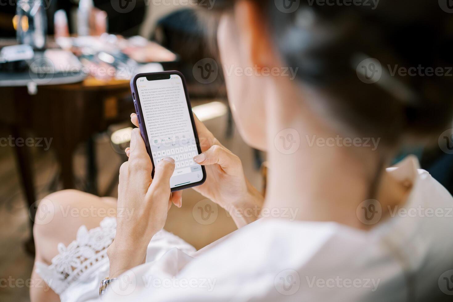 Braut im ein Spitze Kleid sitzt auf ein Stuhl und Typen auf ein Smartphone. zurück Aussicht foto