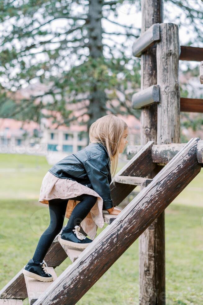 wenig Mädchen klettert das hölzern Treppe auf das Spielplatz im das Park. Seite Aussicht foto