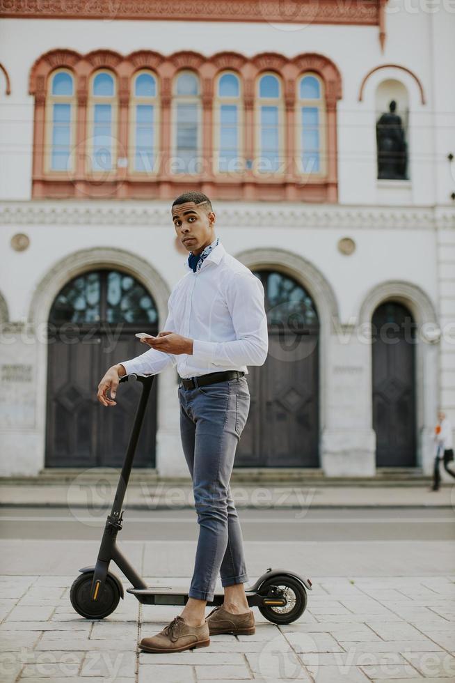 junger Afroamerikaner mit Handy beim Stehen mit Elektroroller auf einer Straße foto