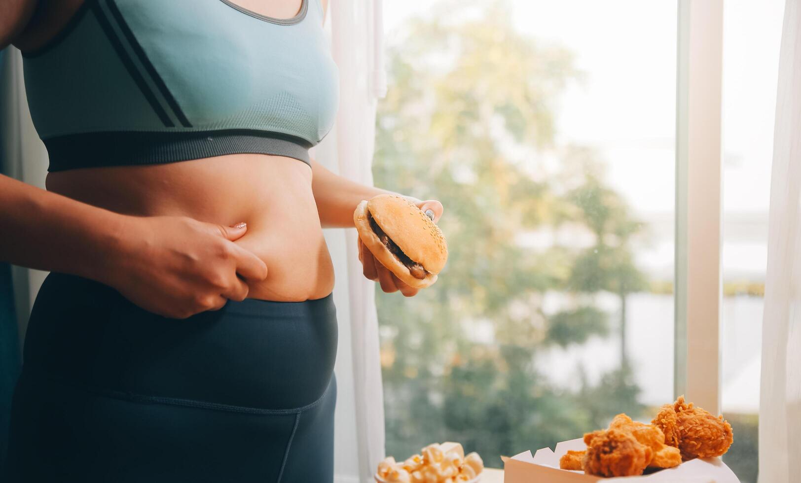 Porträt von fettleibig Frau sieht aus traurig während Kneifen ihr Bauch Fett umgeben durch Müll Lebensmittel. Diät Scheitern Konzept foto