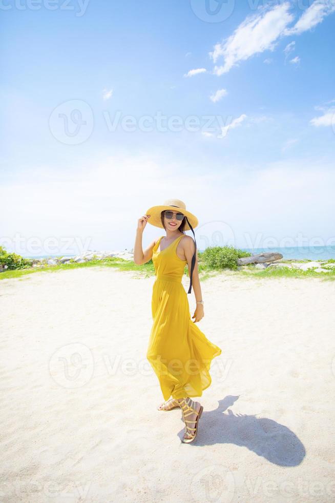 schöne asiatische frau, die gelbe kleidung trägt, die am meerstrand gegen das mittagssonnelicht steht foto