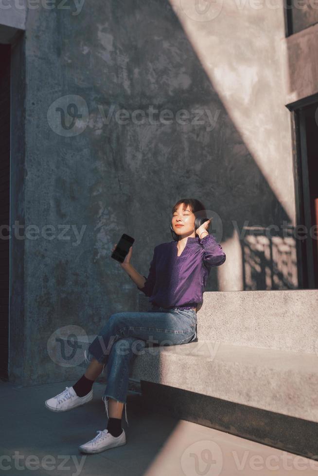 glückliche junge asiatische frau, die musik mit kopfhörern über smartphone hört und spaß hat, während sie auf der straße sitzt foto