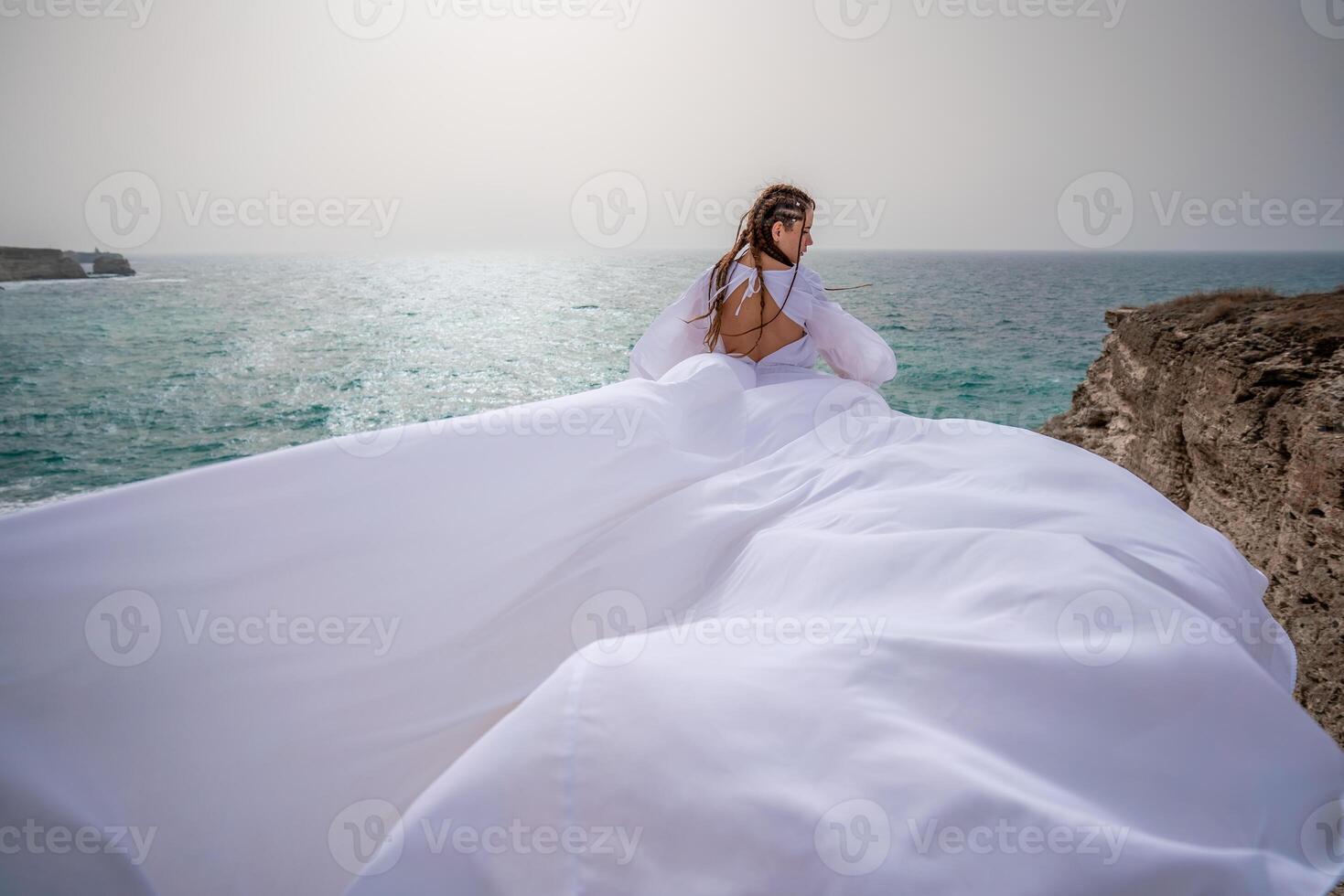 glücklich Freiheit Frau auf das Strand genießen und posieren im Weiß Kleid. Rückseite Aussicht von ein Mädchen im ein flattern Weiß Kleid im das Wind. Feiertage, Ferien beim Meer. foto