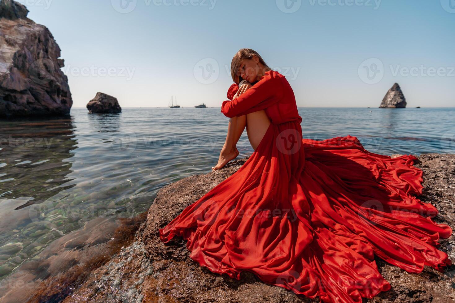 schön sinnlich Frau im ein fliegend rot Kleid und lange Haar, Sitzung auf ein Felsen über das schön Meer im ein groß Bucht. foto