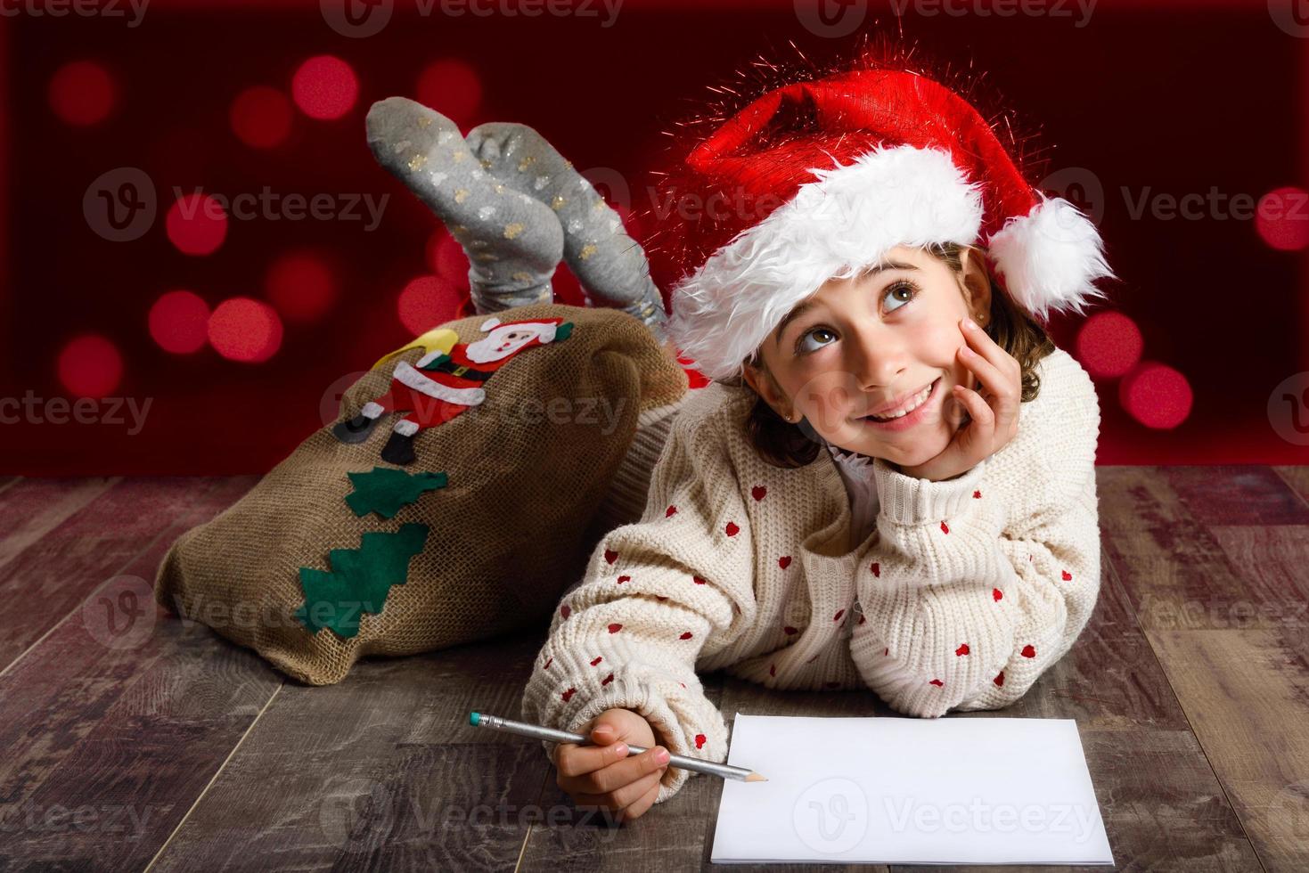 entzückendes kleines Mädchen mit Weihnachtsmütze, das einen Weihnachtsbrief schreibt foto