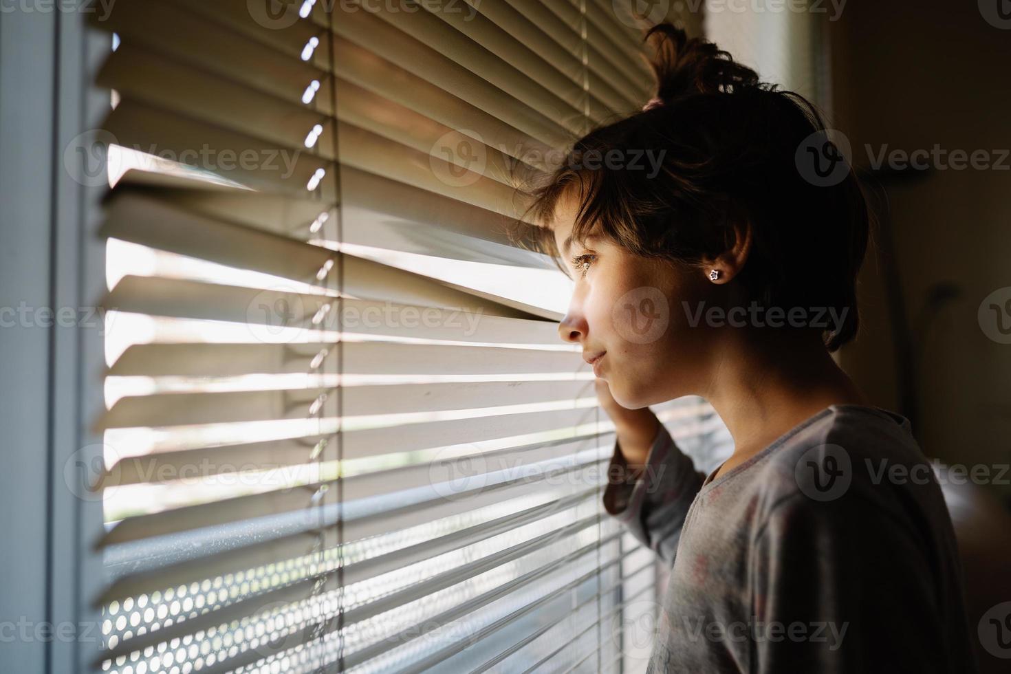 neunjähriges Mädchen, das durch die Jalousien aus dem Fenster schaut foto