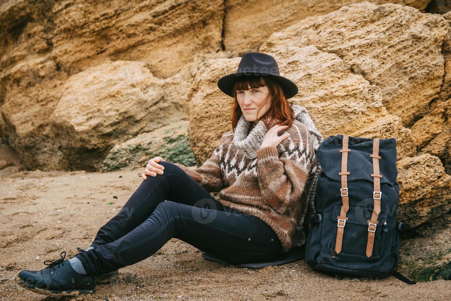 Frau mit Hut und Schal mit Rucksack sitzt auf dem Hintergrund von Felsen foto