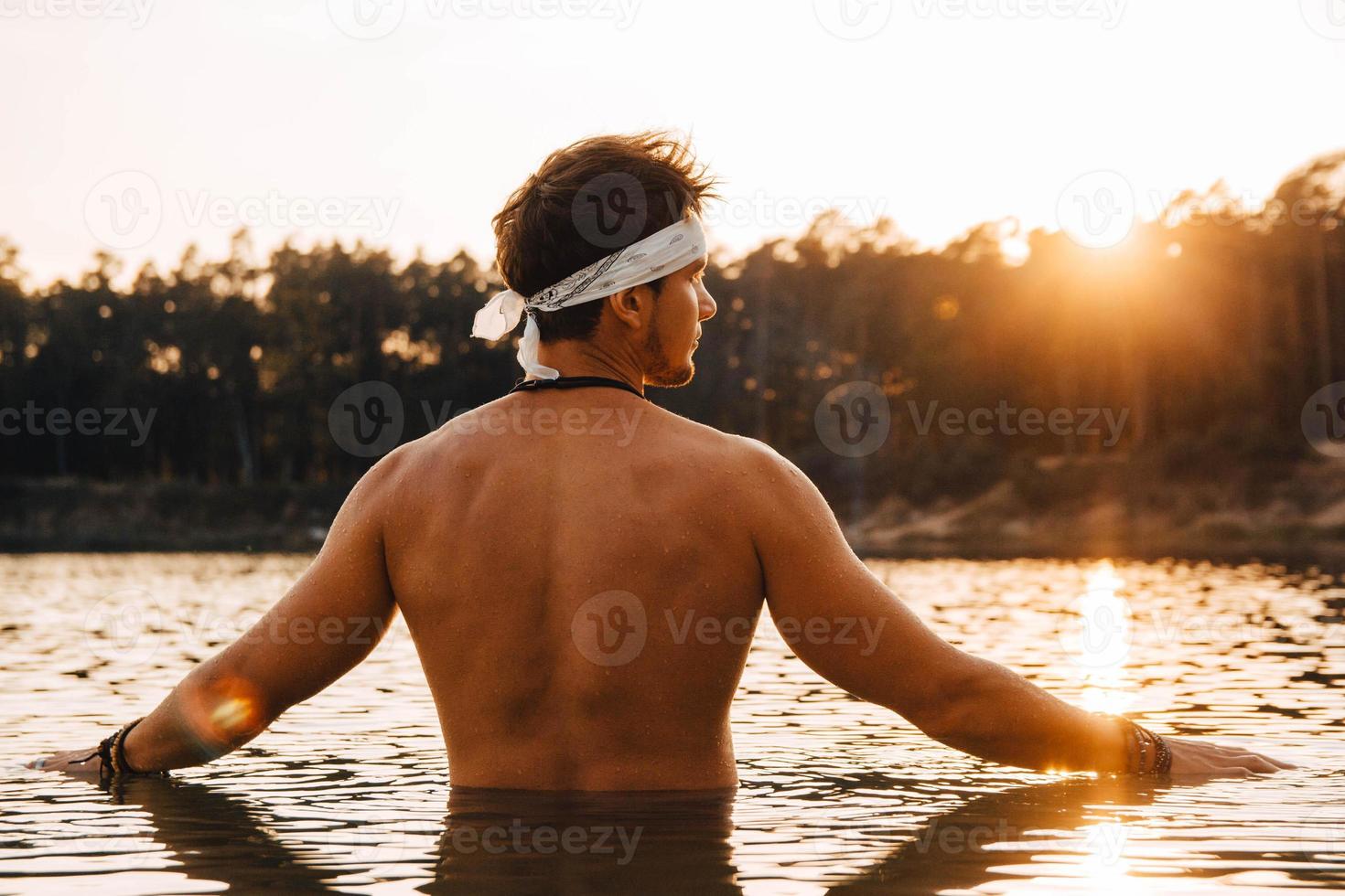 Mann mit perfekter Muskulatur steht bei Sonnenuntergang im Wasser und wirft seine Hände zur Seite foto