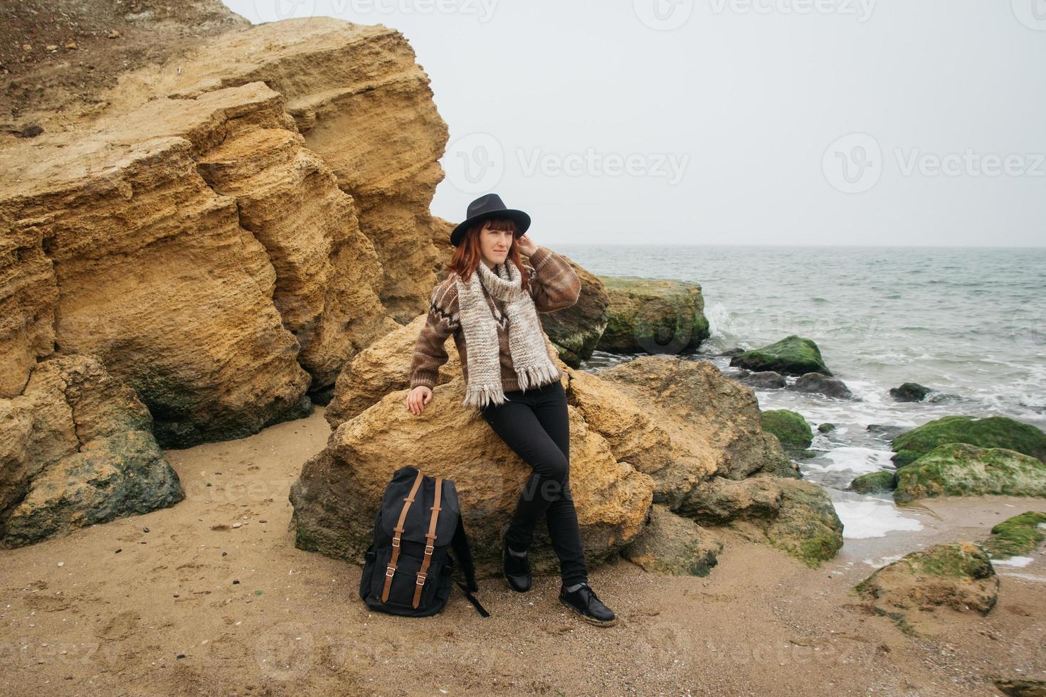 Frau mit Hut und Schal mit Rucksack vor dem Hintergrund von Felsen und schönem Meer foto