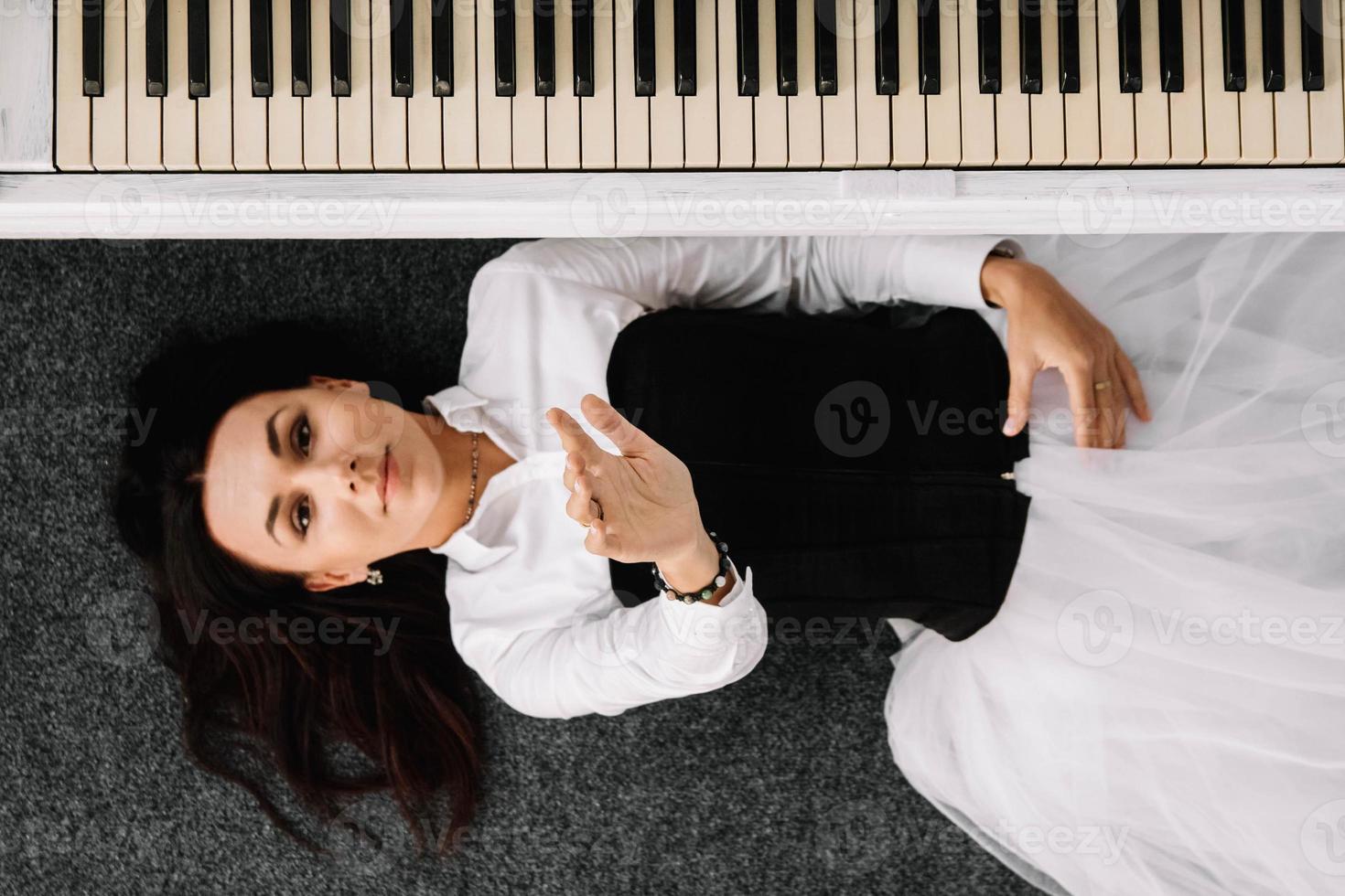 Schöne Frau, die in einem weißen Kleid mit einem schwarzen Korsett gekleidet ist, liegt auf dem Boden in der Nähe des weißen Klaviers. Platz für Text oder Werbung. Sicht von oben foto