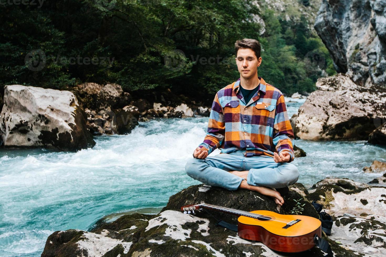 Mann in meditativer Position mit Gitarre sitzt am Ufer eines Bergflusses auf einem Hintergrund von Felsen und Wald foto