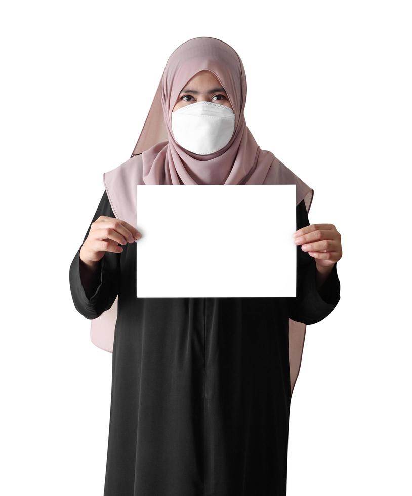muslimische frau mit chirurgischer maske, die leeres papier auf weißem hintergrund hält foto