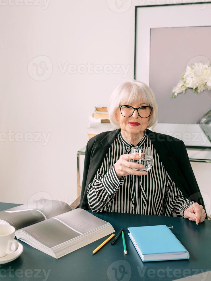 Senior schöne graue Haare Frau Trinkwasser während der Arbeit im Büro. Arbeit, Senioren, Wasserhaushalt, Lösung finden, Konzept erleben foto