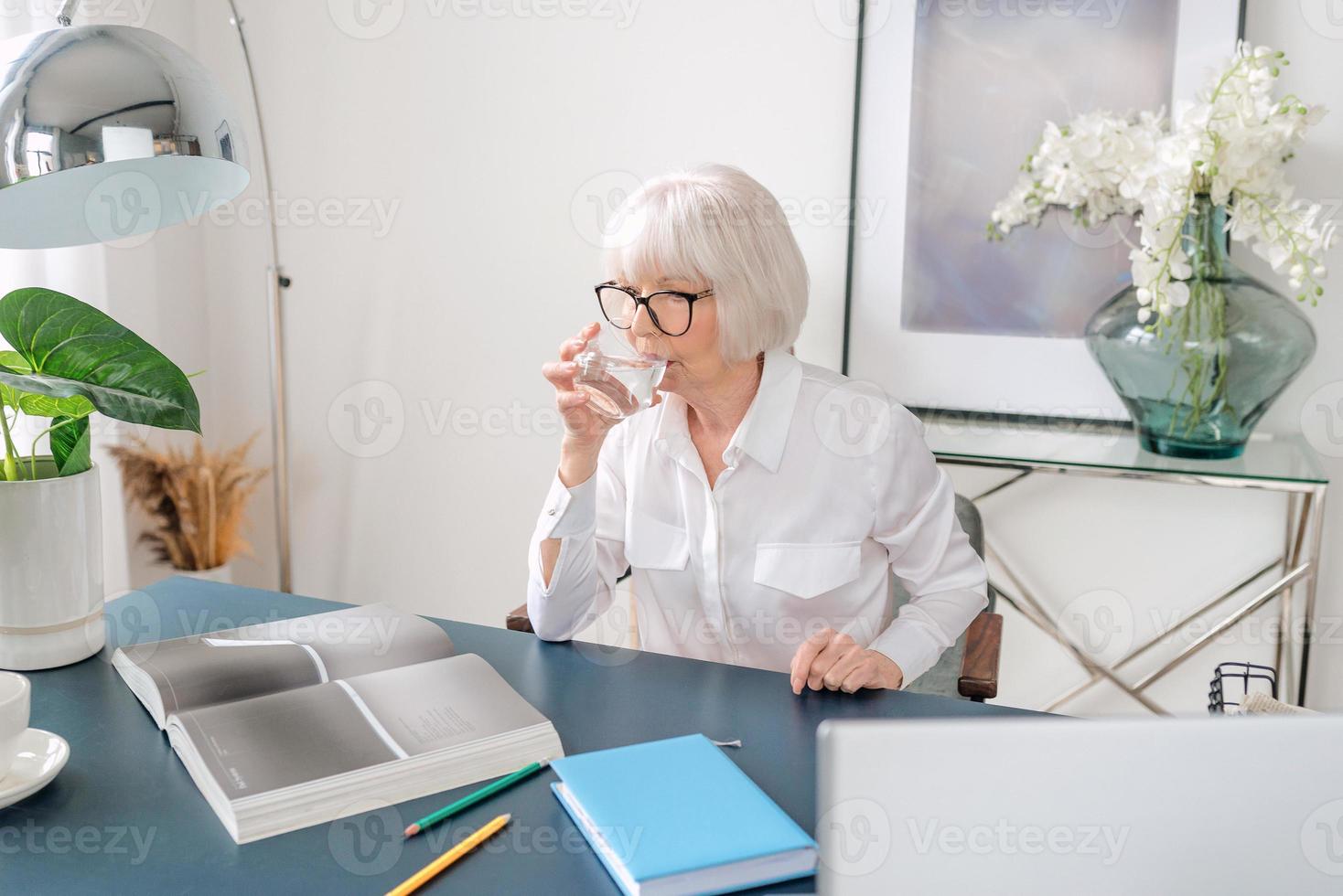 Senior schöne graue Haare Frau in weißer Bluse Trinkwasser während der Arbeit im Büro. Arbeit, Senioren, Wasserhaushalt, Lösung finden, Konzept erleben foto