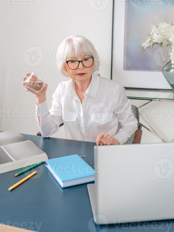 Senior schöne graue Haare Frau in weißer Bluse Trinkwasser während der Arbeit im Büro. Arbeit, Senioren, Wasserhaushalt, Lösung finden, Konzept erleben foto