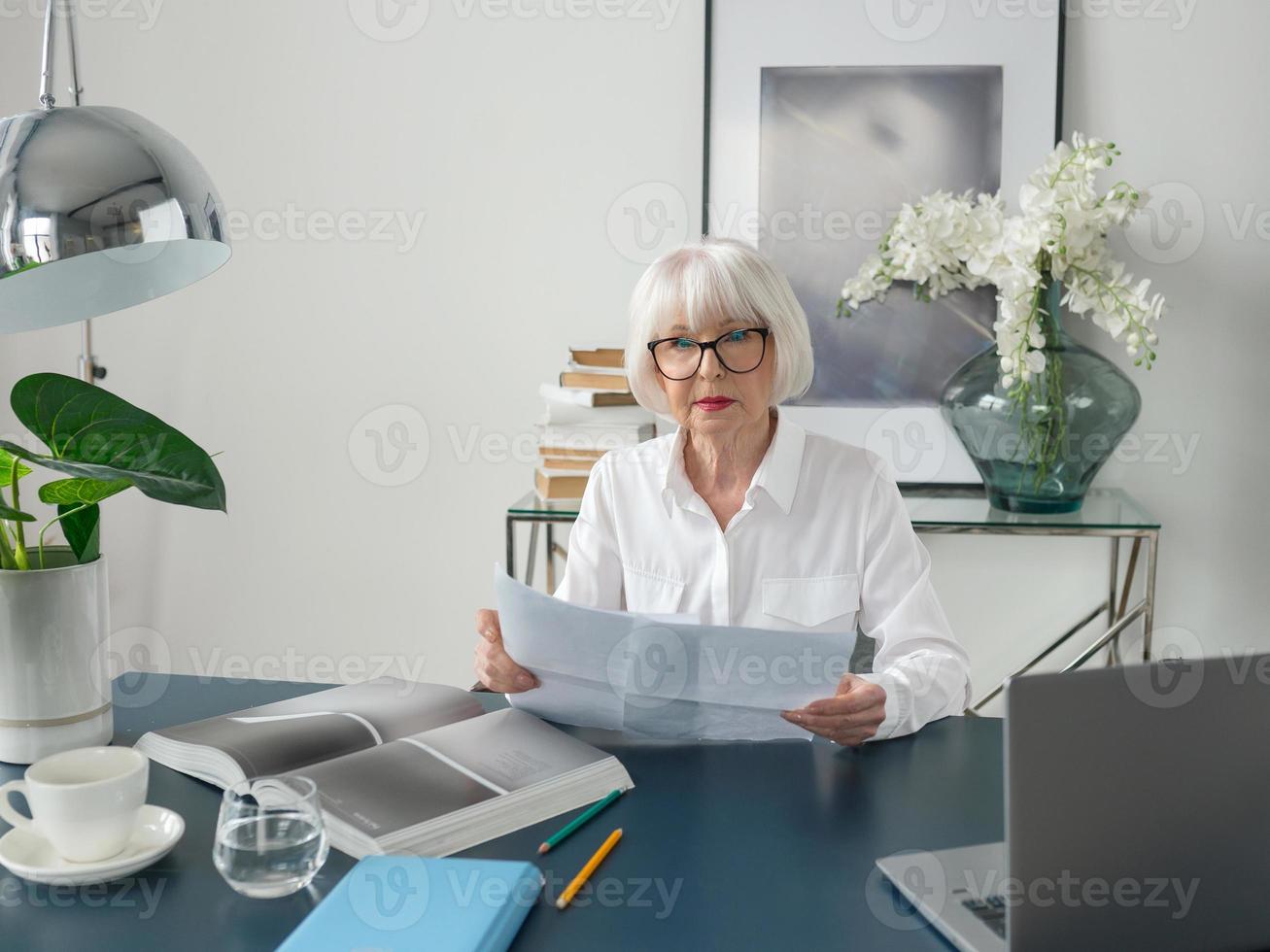 müde senior schöne graue haare frau in weißer bluse lesen dokumente im büro. Arbeit, Senioren, Probleme, Lösung finden, Konzept erleben foto