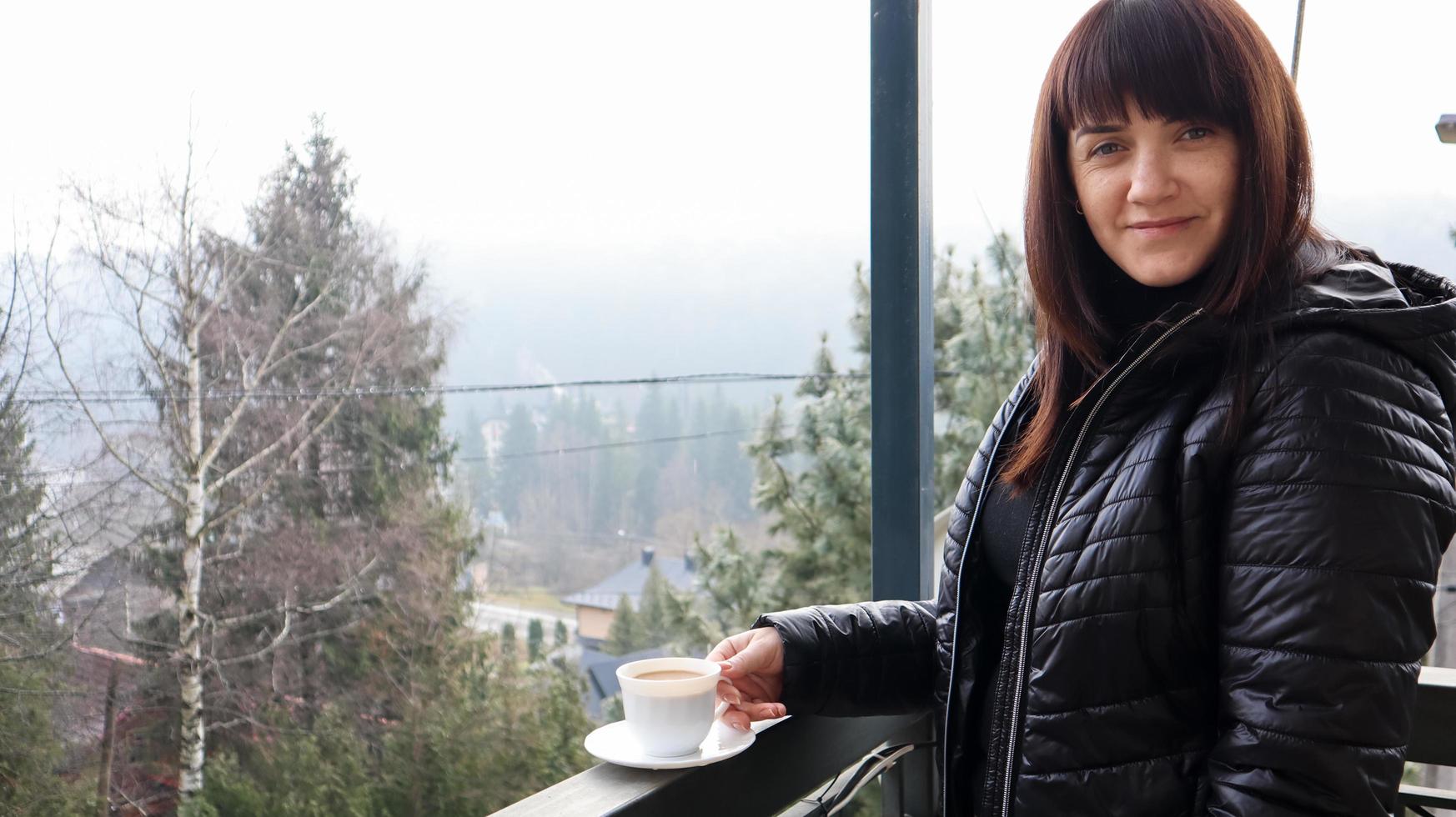 schöne glückliche frau mit einer weißen tasse kaffee oder tee, die den schönen blick auf die berglandschaft im morgennebel vom balkon ihres hotelzimmers genießt. Herbst- und Winterreisekonzept foto