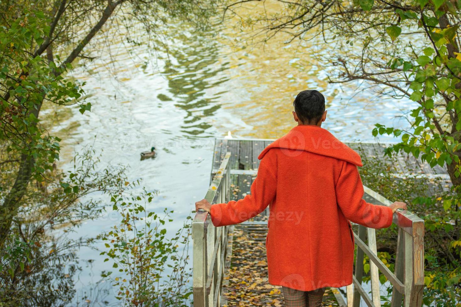 Frau auf dem Rücken mit orangefarbenem Mantel, die zum Flusspier hinuntergeht foto